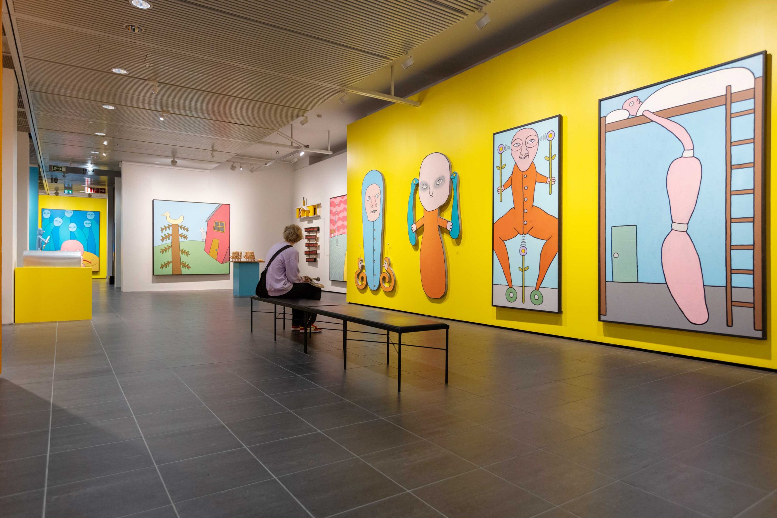 Nuori skeittari istuu taidemuseon näyttelytilassa penkillä ja katselee värikkäitä teoksia, joissa on pelkistettyjä hahmoja.