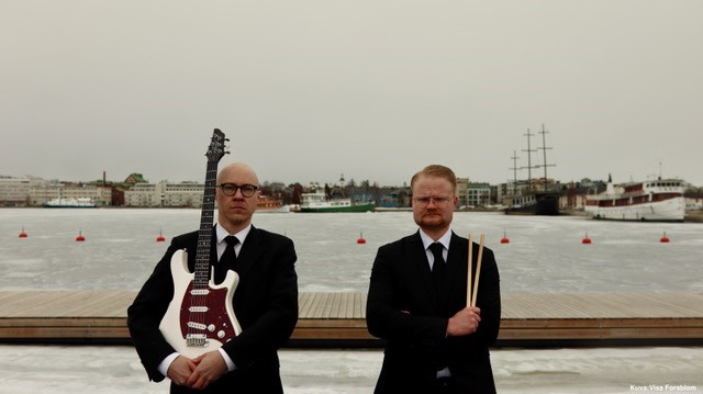 Kaksi miestä seisoo satamassa puvut päällä. Toisella on kädessä kitara ja toisella rumpukapulat.